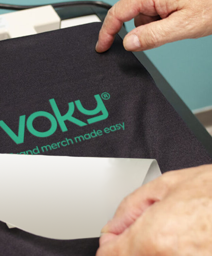 Händer drar bort skydd från textil med Voky-logga och slogan vid tryckpress